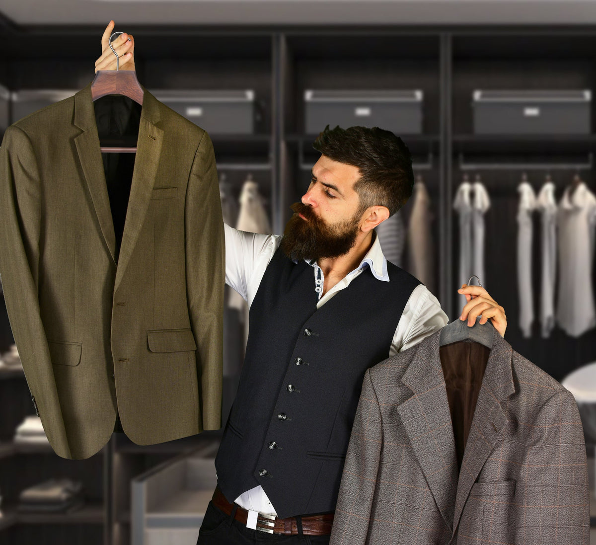 A bearded man in a dark vest, white shirt, and black pants stands in a clothing store holding up three blazers: an olive green one on the left, a gray plaid one on the right, and a dark navy one in the center.